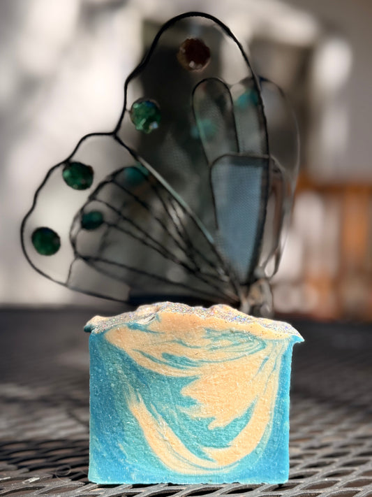 Metal Butterfly-shaped next to a aqua blue and cream Fresh Snow goat milk soap on a metal surface with a blurred background
