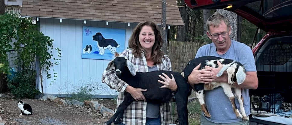 MaryAnn and Hubby each holding young goatsin front of a building with a dog painting on the wall. Cat in background 