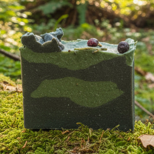 Balsam and spice with junipberries on top of goat milk soap.  colors are green and dark green camo color sitting on moss in a forest.