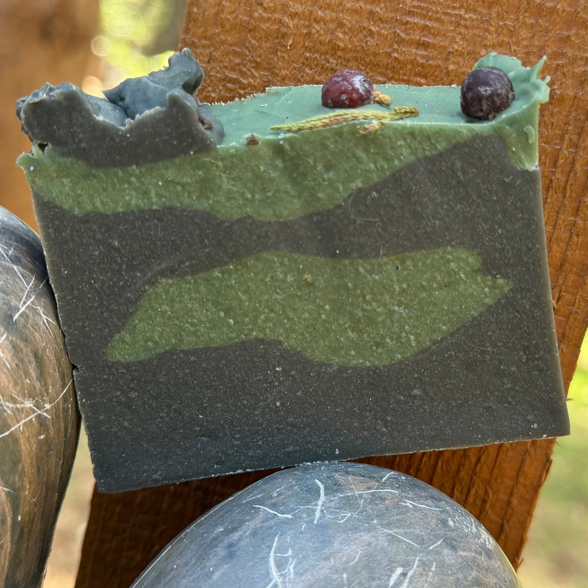 Bar of Balsam & spice goat milk soap with a camouflage pattern pf green and darker green and juniberries on top of soap. on a wooden block outdoors