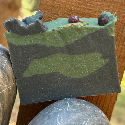 Bar of Balsam & spice goat milk soap with a camouflage pattern pf green and darker green and juniberries on top of soap. on a wooden block outdoors