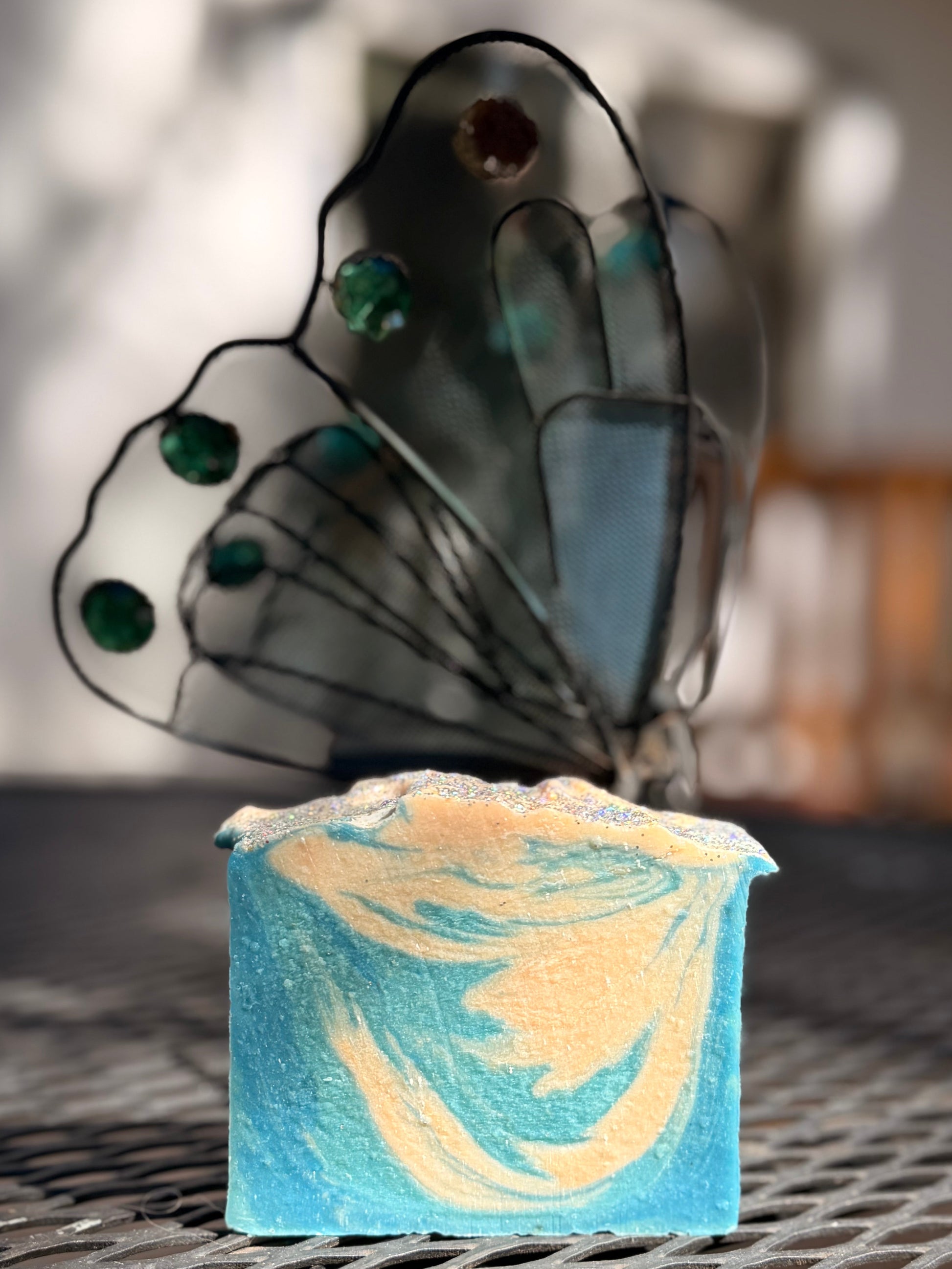 Metal Butterfly-shaped  next to a aqua blue and cream Fresh Snow goat milk soap on a metal surface with a blurred background