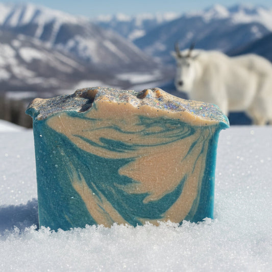 Bar of Fresh Snow goat milk soap with blue and beige swirls in the snow, with mountains and a  mountain goat in the background