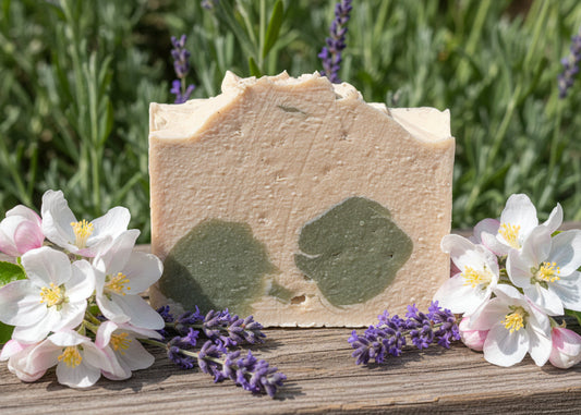 Apple blossom and lavender goat milk soap Bar with green speckles surrounded by flowers and lavender on a wooden surface/