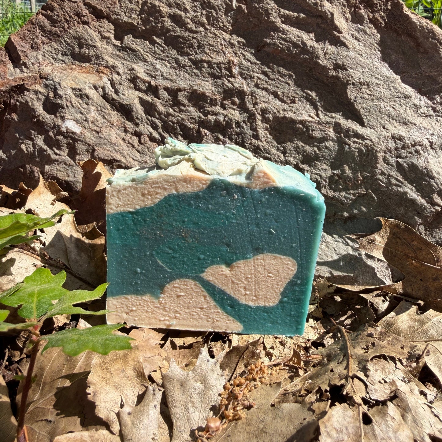 Small square goat milk soap  turquoise and cream goat milk soap called Hawaiian coconut.  Sitting on a  larger rock with leaves and plants around