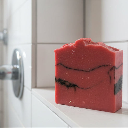 Red soap bar with black streaks on a white surface in a shower - Unscented red clay charcoal goat milk soap.