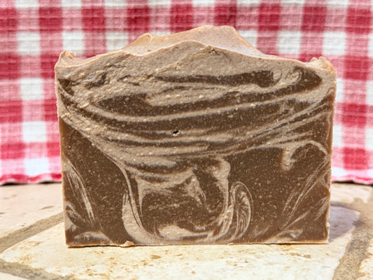 Brown and cream colored swirled cinnamon sugar goat milk soap bar on a stone surface with a red and white checkered cloth in the background.
