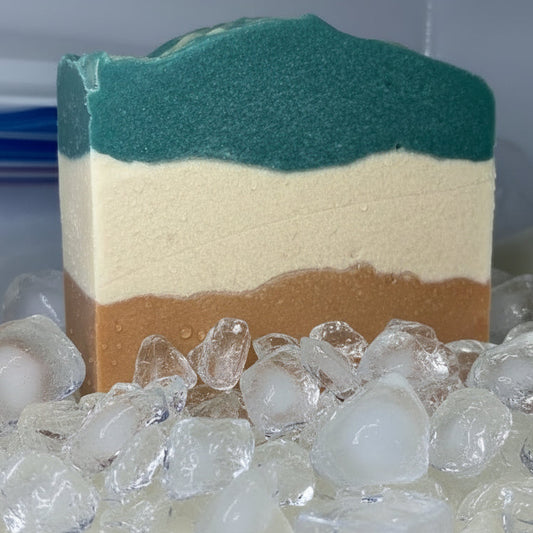 Three-layered popsicle goat milk soap bar with green, white, and brown colors on a bed of ice cubes.